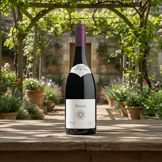 Wine bottle with 'Laurent Miquel Solas' label on a white background. The wine is sat on a wooden table in a garden with vines in the background.