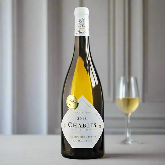 Bottle of 2018 chablis a french white wine. It has a classic white label with gold and black writing. In the background is a glass of wine and grey panelling