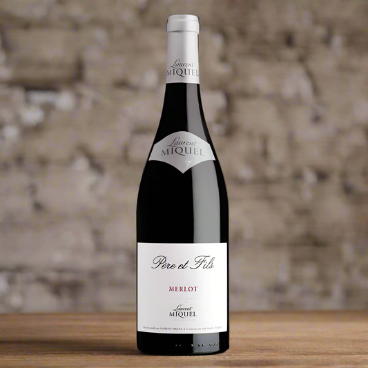 Bottle of red wine with 'Laurent Miquel' written on a white label. Merlot is written in red. The background is a brick wall and the bottle is sat on a wooden table 